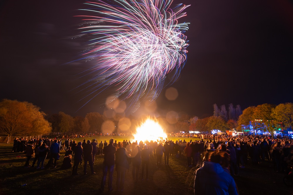 A gathering of people around a large bonfire while fireworks go off overhead.