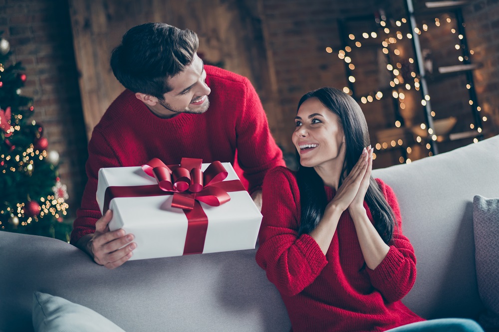 in a festively decorated room, a man hands a present with a bow on it to a woman.