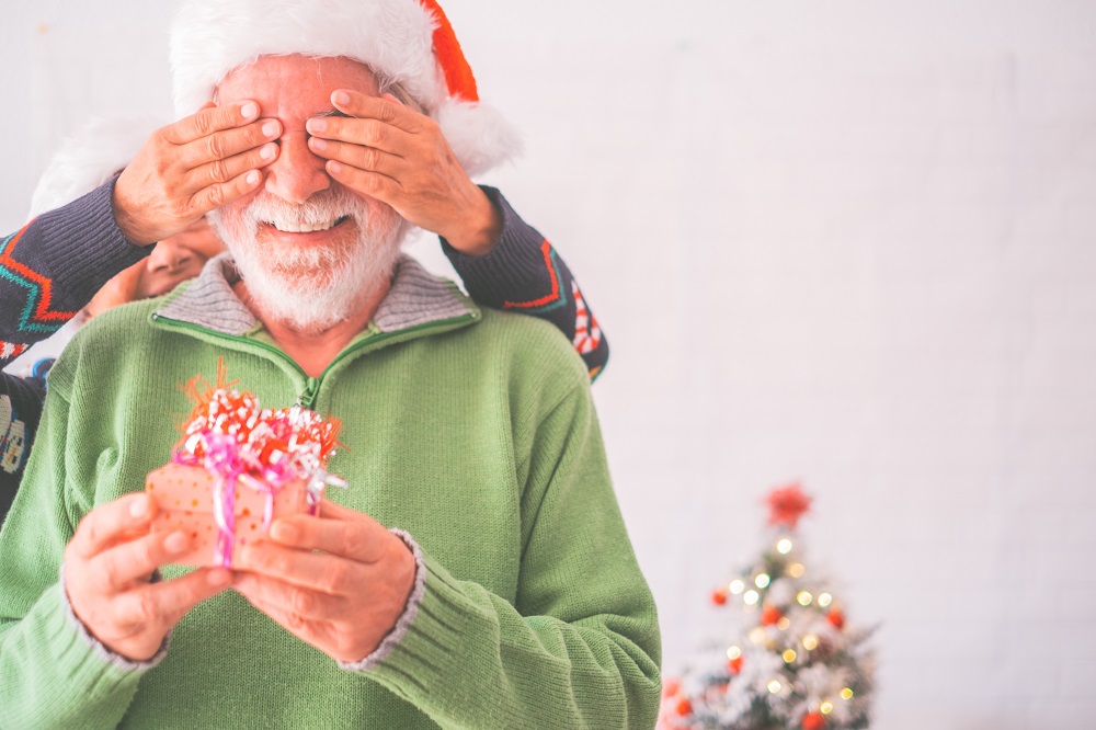 a man wearing a Santa hat holds a boxed gift while a woman covers his eyes with her hands.