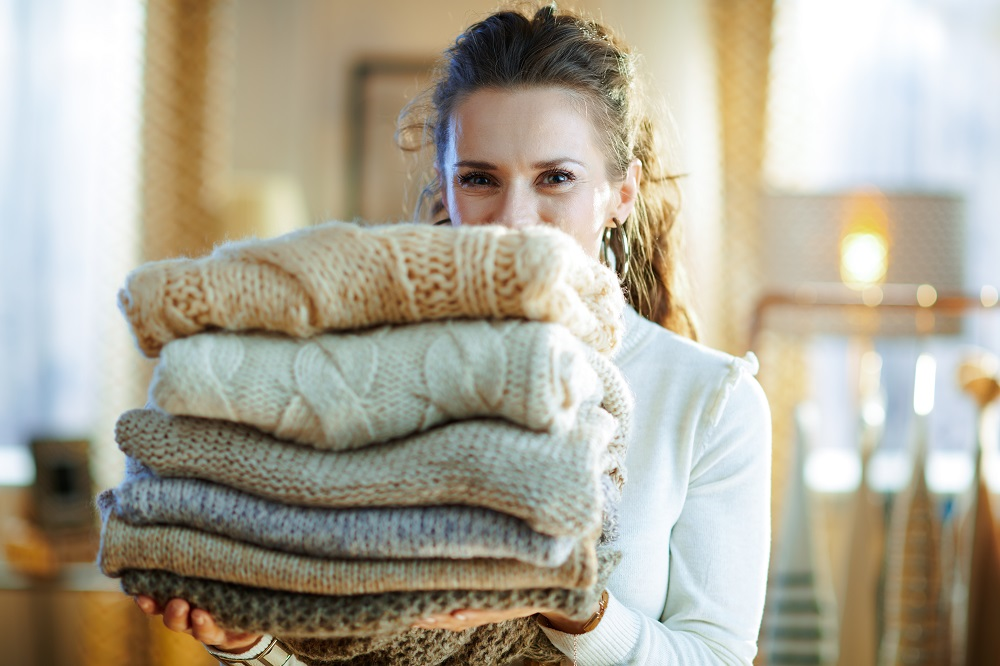 A smiling woman holds a stack of folded woollen jumpers.