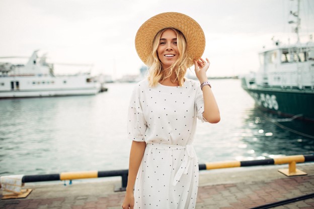 a woman in a summer dress and sunhat stands on a marina.